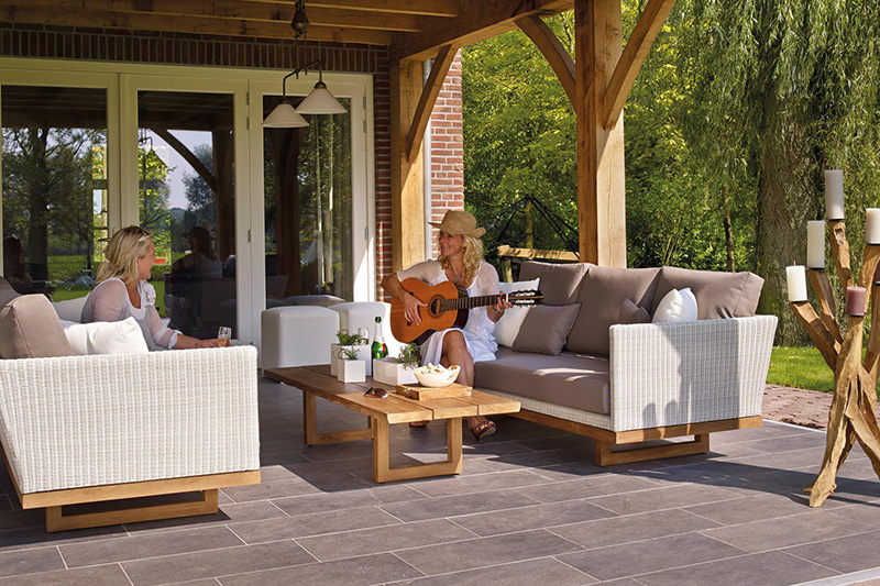 Woman playing guitar sitting on outdoor sofa on patio covered by pergola roof.